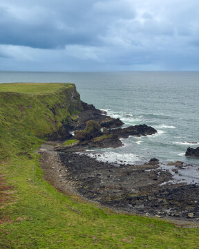 Green cliffs descend to a rugged volcanic shoreline beneath stormy clouds near Giant's Causeway on the Causeway Coast of Northern Ireland.