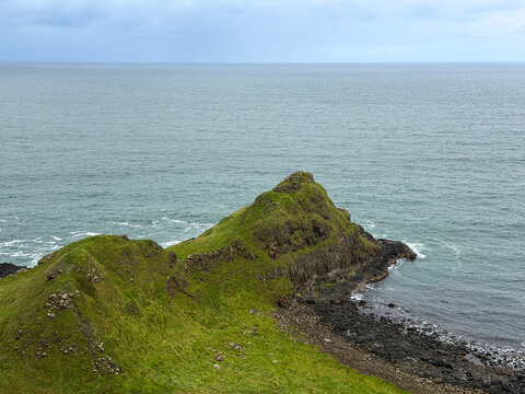 A rugged green headland rises above dark volcanic rocks and the North Atlantic along the scenic Causeway Coast near Giant's Causeway, Northern Ireland.