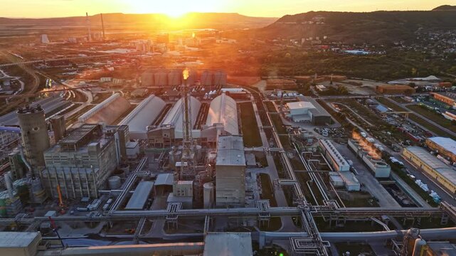 The sun sets over an industrial area filled with factories and pipelines. Smoke rises from the structures as the landscape extends into the valley and hills.