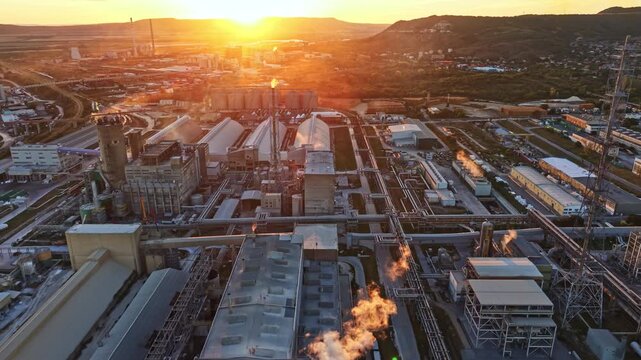 Factories and industrial buildings are seen under a sunset with smoke rising into the sky. The scene shows a busy area that is part of a larger city.