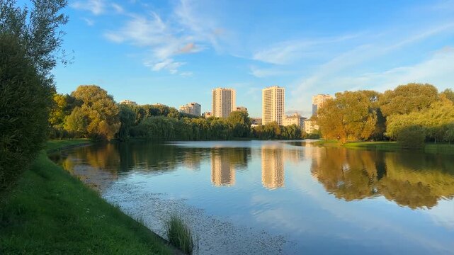 Lake in city park with trees. Tall buildings rise background. Residential buildings near lake. Scene in city park. Reflections in park pond with multifamily apartments. Long panoramic river downtown