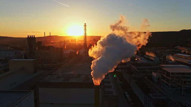 Smoke billows from a factory in an industrial zone as the sun sets in the background. The scene shows buildings and structures silhouetted against the sky.
