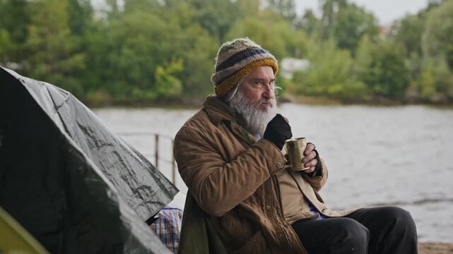 Senior Caucasian homeless man sitting near tent campsite, eating canned food with spoon. Ideal for charity projects, social awareness, and documentary editing