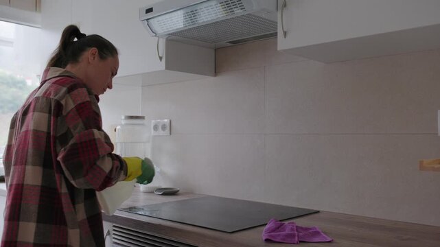 Woman in protective rubber gloves sprays detergent and wipes an induction stove surface with a microfiber cloth in a modern kitchen.