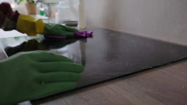 Close-up of a person wearing protective rubber gloves wiping and cleaning a black ceramic induction stovetop with a microfiber rag in a home kitchen.