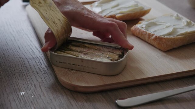 Close-up of a person opening a tin can of preserved fish on a wooden cutting board next to bread with butter in a kitchen.