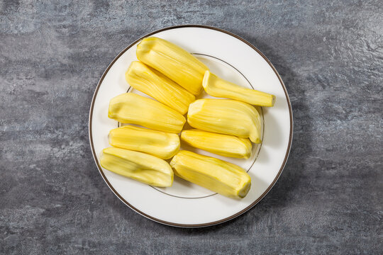 A top-down view of peeled, ripe yellow jackfruit pieces arranged on a white plate with a thin brown rim, placed on a gray textured surface.