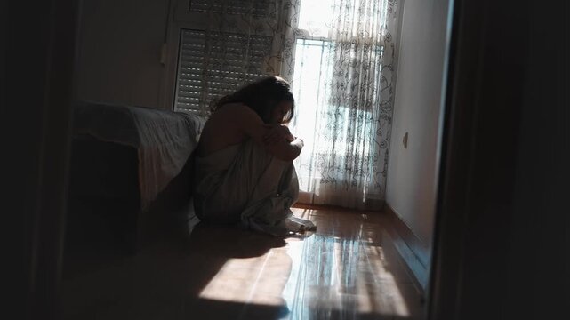 Depressed young woman sitting on the floor beside her bed wrapped in a bedsheet near a window, experiencing sadness, loneliness and emotional distress at home.
