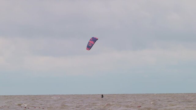 Action wide shot of a lone kitesurfer with kite gliding across rough ocean waters on an overcast day. Extreme water sports and active lifestyle concept. Flying over water in slow motion..