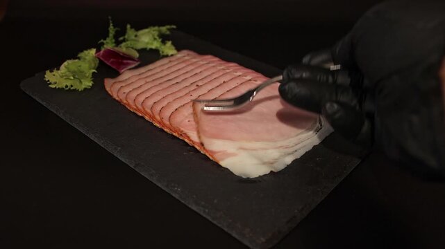 Close-up of chef in black gloves taking a slice of smoked ham with a fork on slate plate. Perfect stock footage for culinary ads, charcuterie videos, and food menus.