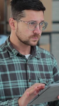 Vertical video. Worker checking inventory, writing notes on tablet. A man works in a warehouse with rows of shelves with boxes, packages.