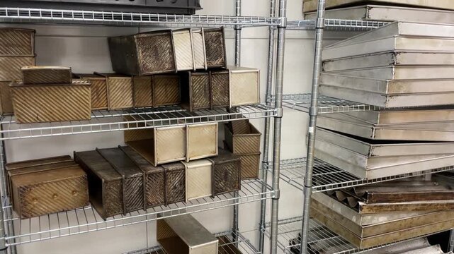 organized storage scene with stacked ribbed loaf pans and baking sheets on chrome wire racks inside commercial bakery room, showing worn metal surfaces and patina