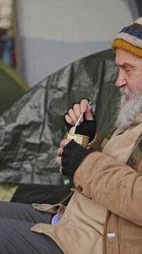 Vertical video of senior Caucasian homeless man eating canned food with plastic spoon near street shelter. Clip fits charity campaigns and poverty awareness videos