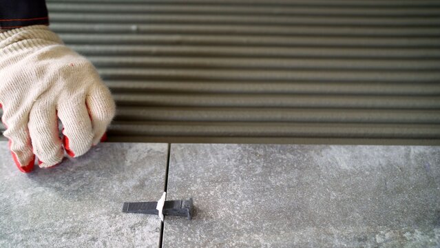 Worker in protective gloves laying large grey ceramic tiles on floor with adhesive and leveling system during home renovation