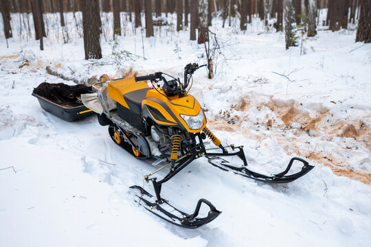 Compact orange snowmobile with towing cargo sled in winter forest. Utility snowmobile with drag sled for hauling gear. Winter transport for hunting and fishing.
