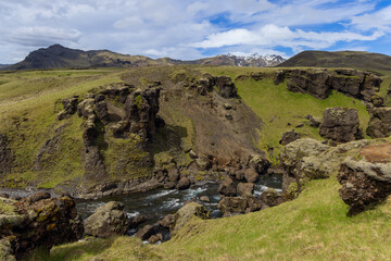 Obraz premium Skoga River flows between dark rock walls and scattered boulders in a grassy Iceland gorge, with rugged ridges and distant snow patches beneath a partly cloudy blue sky.