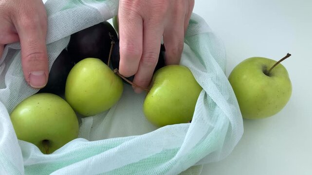 A woman puts apple and plum on a white table from eco bag. A reusable textile net, mesh with juicy apples and plums, a generous summer harvest, the freshness of the fruit, the natural bounty of nature