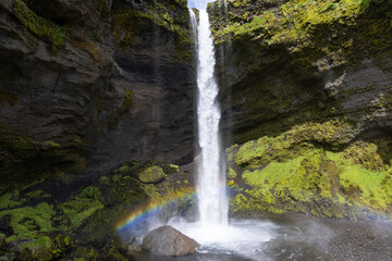 Obraz premium Kvernufoss waterfall drops between layered cliffs in Iceland, with a rainbow crossing spray above the broad plunge pool and bright green vegetation covering the right canyon wall.