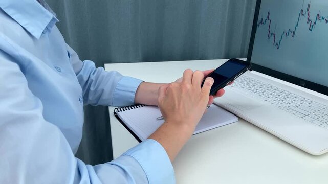 The woman is sitting at a white table, searching for information online, using her phone, assessing market trends using computer charts and records, and planning her further investment strategy
