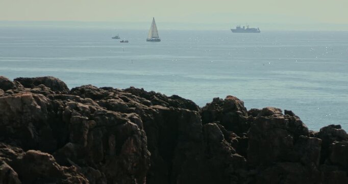 View of Portugal&rsquo;s  Atlantic coastline with dark rocks in the foreground and a variety of vessels &mdash; including a fishing boat, yacht, and ship &mdash; sailing in the brightly sunlit ocean in the background.