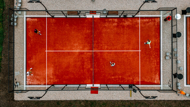 Aerial view of two padel courts with players in action, featuring red clay surfaces, surrounded by fencing and seating areas, showcasing a vibrant sports environment