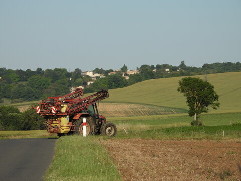 A red tractor with a large sprayer stands beside a narrow road. The nearby field, mixing green grass and brown soil, reflects ongoing agricultural work. 