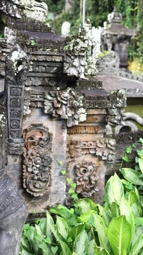 Ornate temple garden stone carvings stand amid lush tropical plants, with weathered shrine reliefs filling frame. Temple garden details feel serene, and carved stone faces and mossy surfaces show age.