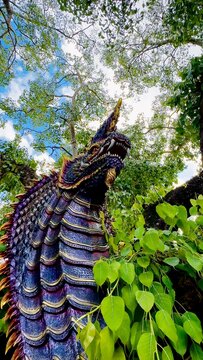 Colorful Naga serpent sculpture beside a large tree in a lush outdoor setting, with intricate scales and vivid details surrounded by green foliage and bright blue sky.