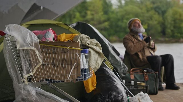 Senior Caucasian homeless man sitting near tent encampment with full shopping cart in foreground. Footage is suitable for social awareness campaigns and poverty documentaries