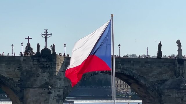 Czech flag flapping in the wind near prague bridge, viewed from a distance with crosses and people visible in the background. the flag red and white colors.