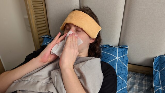 Sick young man with long brown hair lying in bed with wet towel on forehead and blowing his nose with white tissue. Treating seasonal flu or cold at home.
