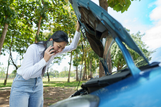 Stressed Asian woman talking on phone by broken car for roadside help