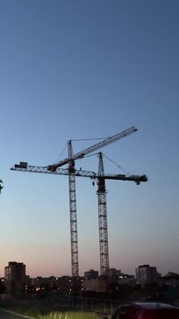 two tall lattice tower cranes stand over urban construction site at dusk with long horizontal jibs crossing near top, suspended hook visible.