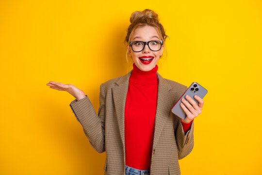 Young woman holding smartphone and raising hand with a surprised smile on bright yellow background.