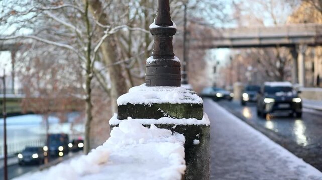 Winter scene in Paris with a close-up of a traditional lamp post covered in snow along a cobblestone street. A car with headlights on is driving in the background