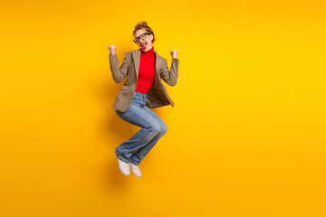 Smiling young woman in trendy jacket and jeans jumps with cheerful gesture on bright yellow...