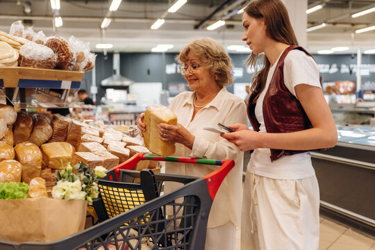 Senior woman with gray hair examines a loaf of bread while shopping with a young woman in a grocery store aisle filled with various types of bread and baked goods
