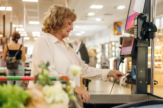 Senior woman with curly hair using self-checkout machine in grocery store, fresh produce and shopping cart visible in the foreground