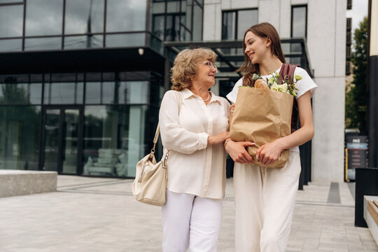 Two women, one elderly and one young, walking together outside a modern building, carrying grocery bags filled with fresh produce and smiling at each other