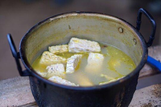 Traditional seasoning process of tempeh with herbs in an old aluminum pot before frying.