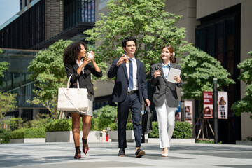 Fototapeta premium A group of diverse professionals walks together outdoors at financial district building, holding coffee cups and talking while heading to work, conveying a sense of teamwork, connection, and ambition