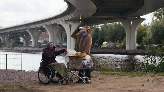 Senior Caucasian homeless man sharing hot drink with companion with disability in wheelchair outdoors. Clip fits charity campaigns and social support projects