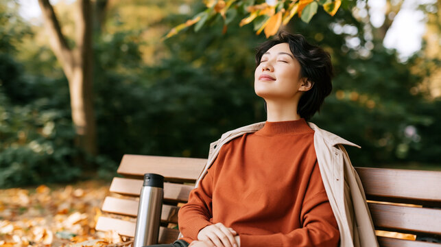 Asian woman sitting on a wooden bench in a park, wearing a rust-colored sweater and light jacket, enjoying a sunny autumn day surrounded by colorful leaves