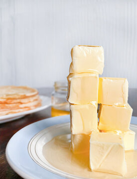 A rustic still life featuring cubes of butter drizzled with honey, with thin pancakes and a jar of honey in the background.