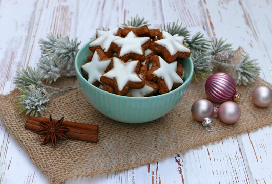 Traditional cinnamon stars in a light green bowl with cinnamon sticks, star anise, and pink Christmas baubles on a white wooden table.