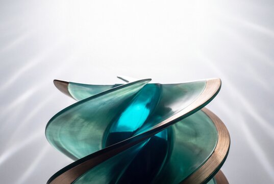 Abstract Spiral Sculpture Made of Translucent Teal Glass and Brushed Bronze Metal Edges on a Bright Background