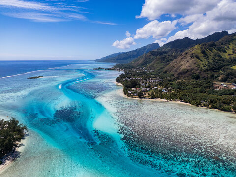 Aerial view of Mo'orea island featuring turquoise lagoon waters, coral reefs, lush green mountains, and coastal vegetation in Moorea-Maiao, Windward Islands, French Polynesia.