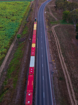 Aerial view of a long freight train traveling on tracks parallel to a highway through rural fields and trees in Home Hill, Queensland, Australia.