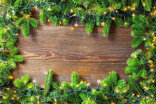 Christmas wooden background with bright fairy lights and fir branches frame.