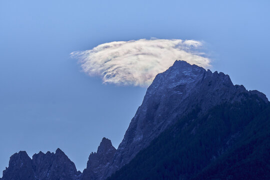 Aerial view of Valchiavenna mountain peak with a rare iridescent lenticular cloud hovering above the rocky summit under a clear blue sky in Lombardy, Italy.